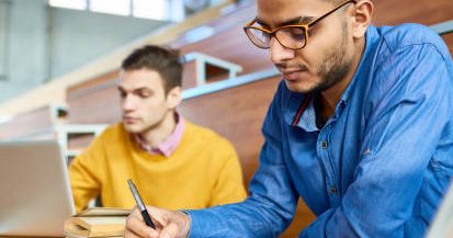 Portrait of two students sitting at desks in modern auditorium at college and preparing for class, focus on young Middle-Eastern man writing in copybook